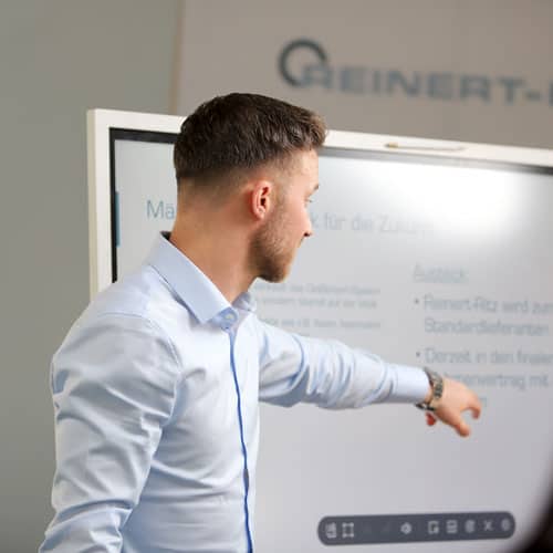 Ein Mann steht mit dem Rücken zu einem Bildschirm und erklärt mit dem Finger einen Zusammenhang, den der Bildschirm zeigt